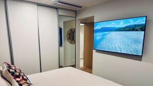 A bedroom with a bed, a colorful pillow, a wall-mounted TV displaying a beach scene, and a mirror. - Bosques de Jacó Condo (Jacó)