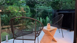 A patio with two wicker chairs, a small wooden table, and a glass enclosure with a view of lush greenery. - Bosques de Jacó Condo (Jacó)