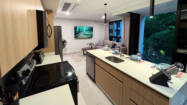 A modern kitchen with a black stove, wooden cabinets, a large island with a sink, and a view of a forest through a window. - Bosques de Jacó Condo (Jacó)