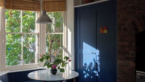 Une pièce avec une baie vitrée, une table ronde ornée d’un vase de fleurs, un meuble bleu et un mur en briques.