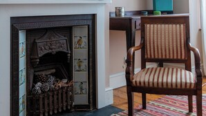 Une pièce avec une cheminée, un grand miroir, une chaise, une table avec une lampe et un tableau accroché au mur.