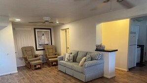 Ein Wohnzimmer mit einem gestreiften Sofa, zwei Sesseln, einem Deckenventilator und einem durch einen offenen Durchgang einsehbaren Küchenbereich.