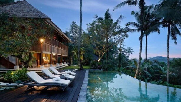 An infinity pool with lounge chairs, a wooden deck, and a house with a thatched roof surrounded by palm trees. - Villa Inka Ubud by Nakula (Bali)