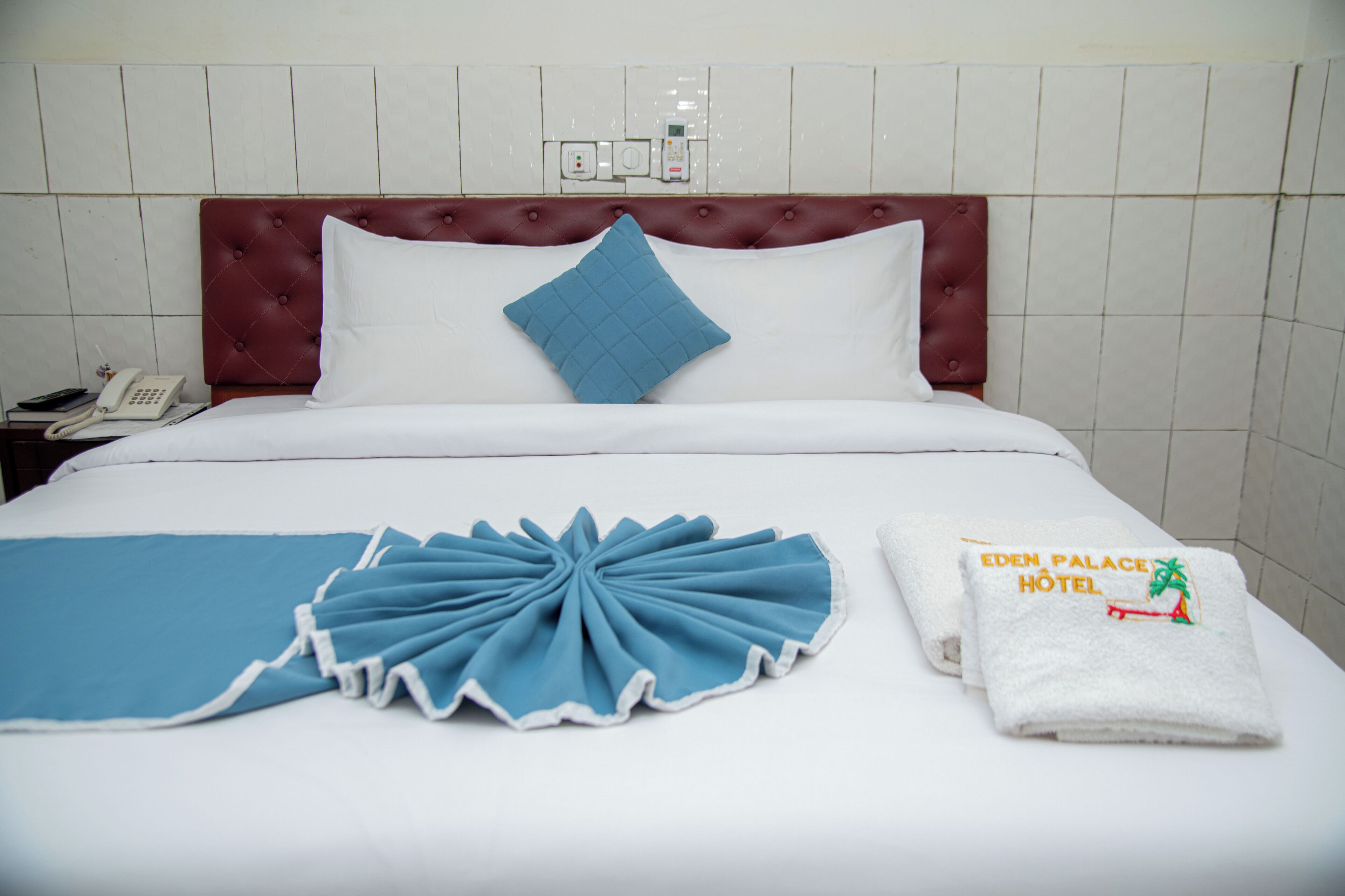 Una cama bien hecha con un cojín decorativo azul, un cojín blanco y una manta azul. Hay una toalla blanca con el texto 'EDEN PALACE HOTEL' y un diseño de palmera.