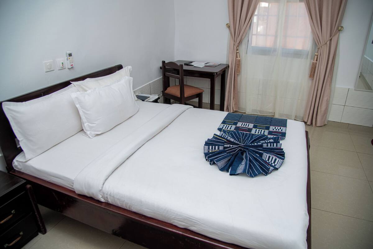 A neatly made bed with white linens, a wooden headboard, and a blue and white patterned towel on the bed. There is a small wooden table with a chair beside the bed, and a window with curtains.