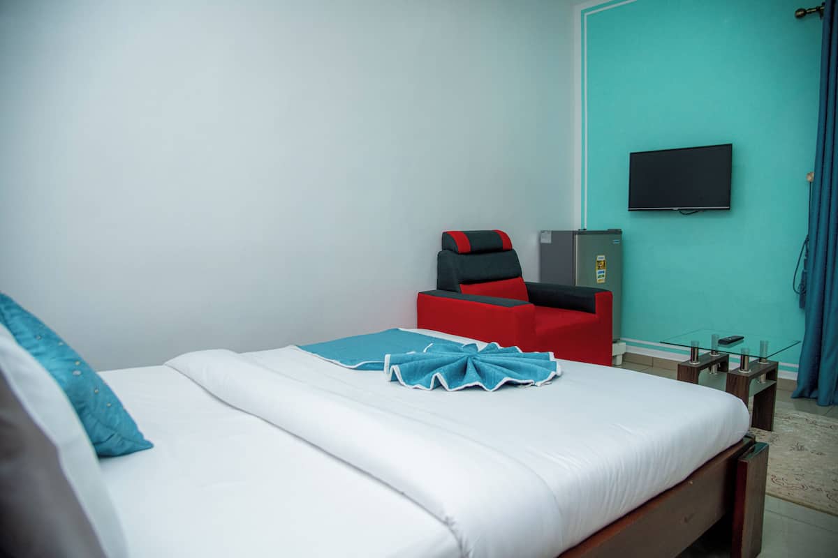 A neatly made bed with white and blue bedding, a red chair, a television on the wall, and a small table with a glass top.