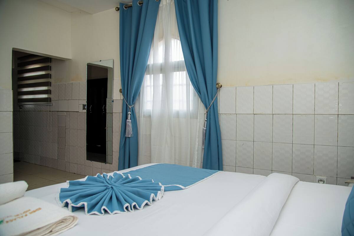 A hotel room with a bed, a bathroom with a shower, and a view of the outside through a window with blue curtains.