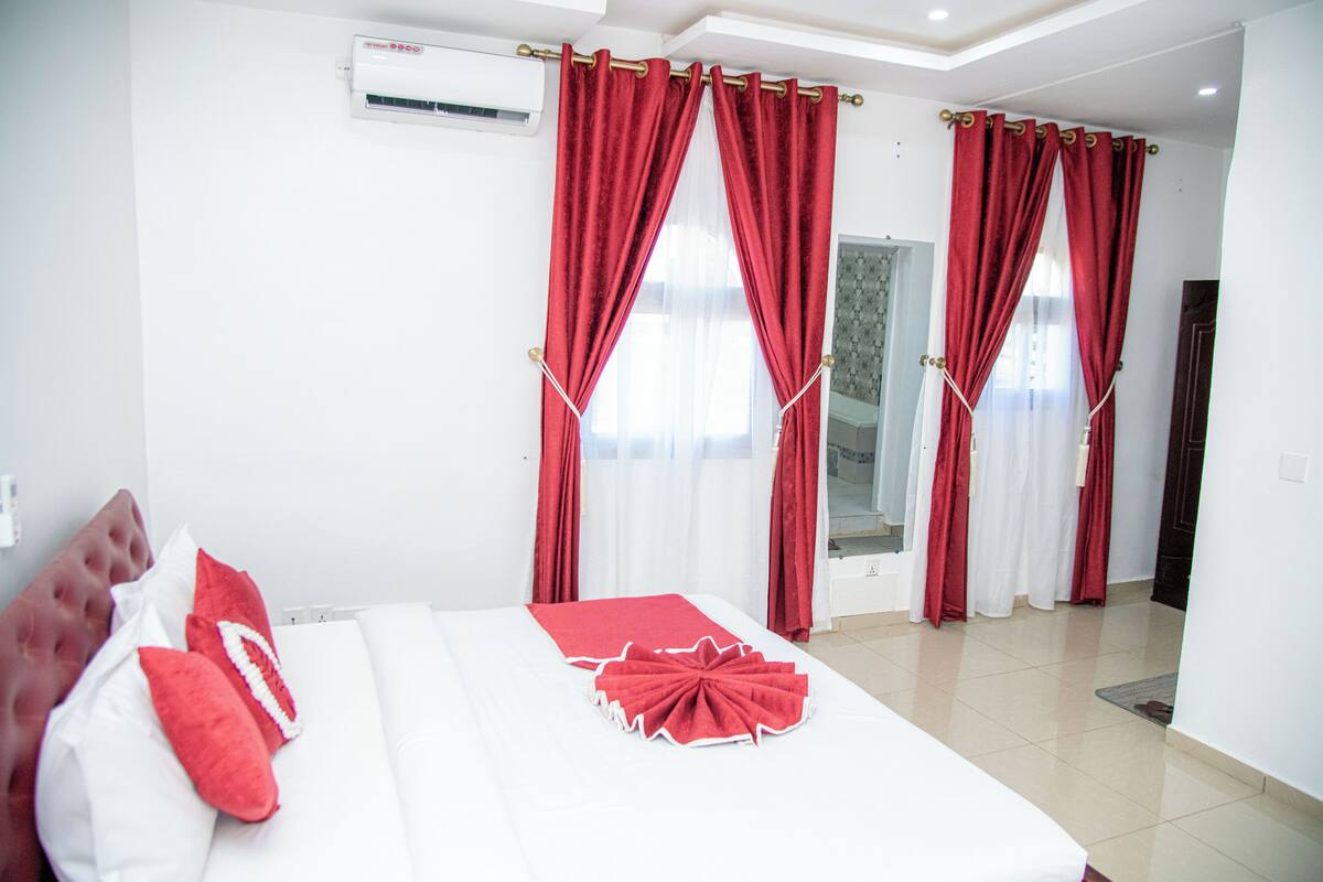 A hotel room with a bed, red curtains, white walls, and a window.
