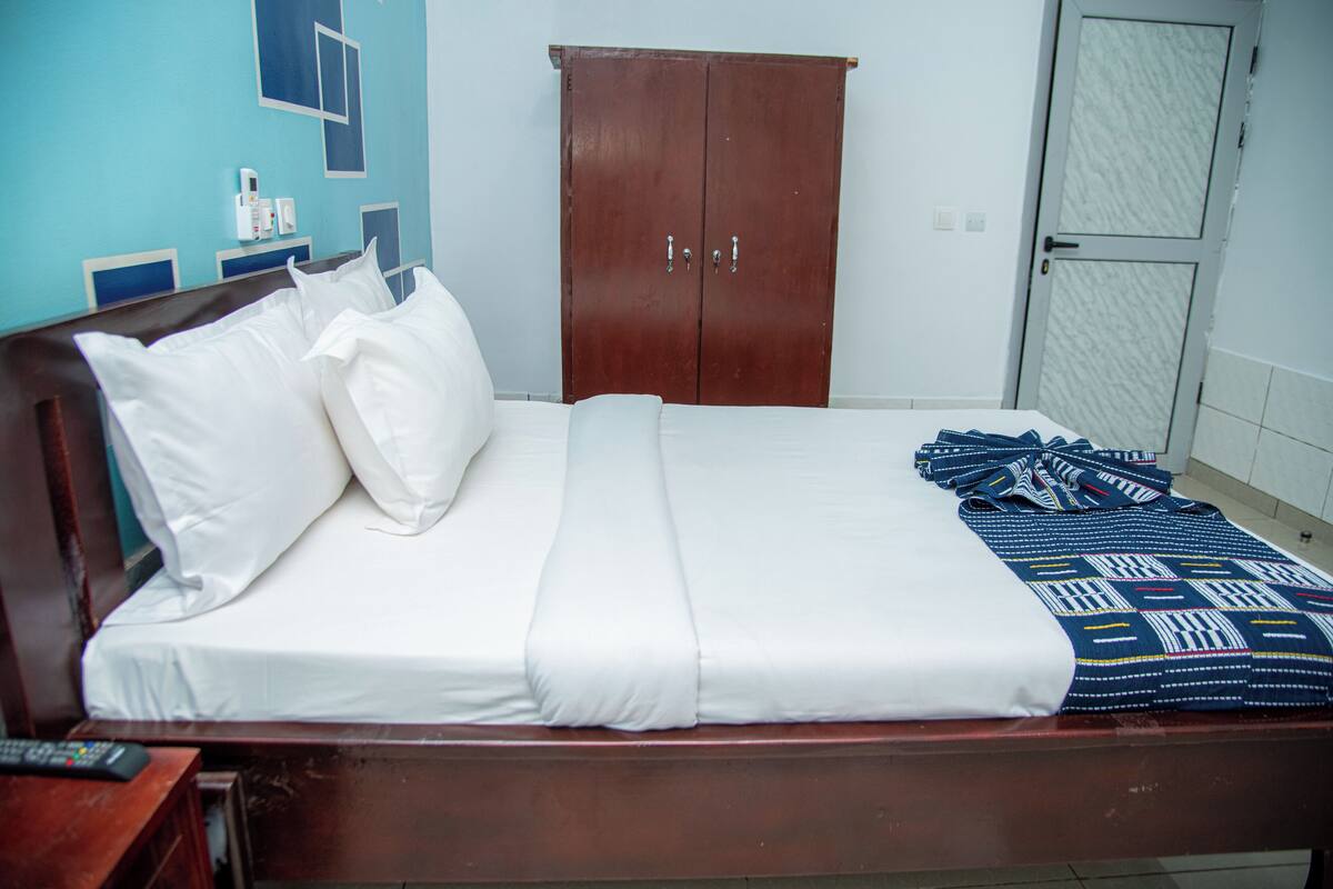 A neatly made bed with white linens and a patterned blanket, a wooden wardrobe, and a small bedside table with a remote control.