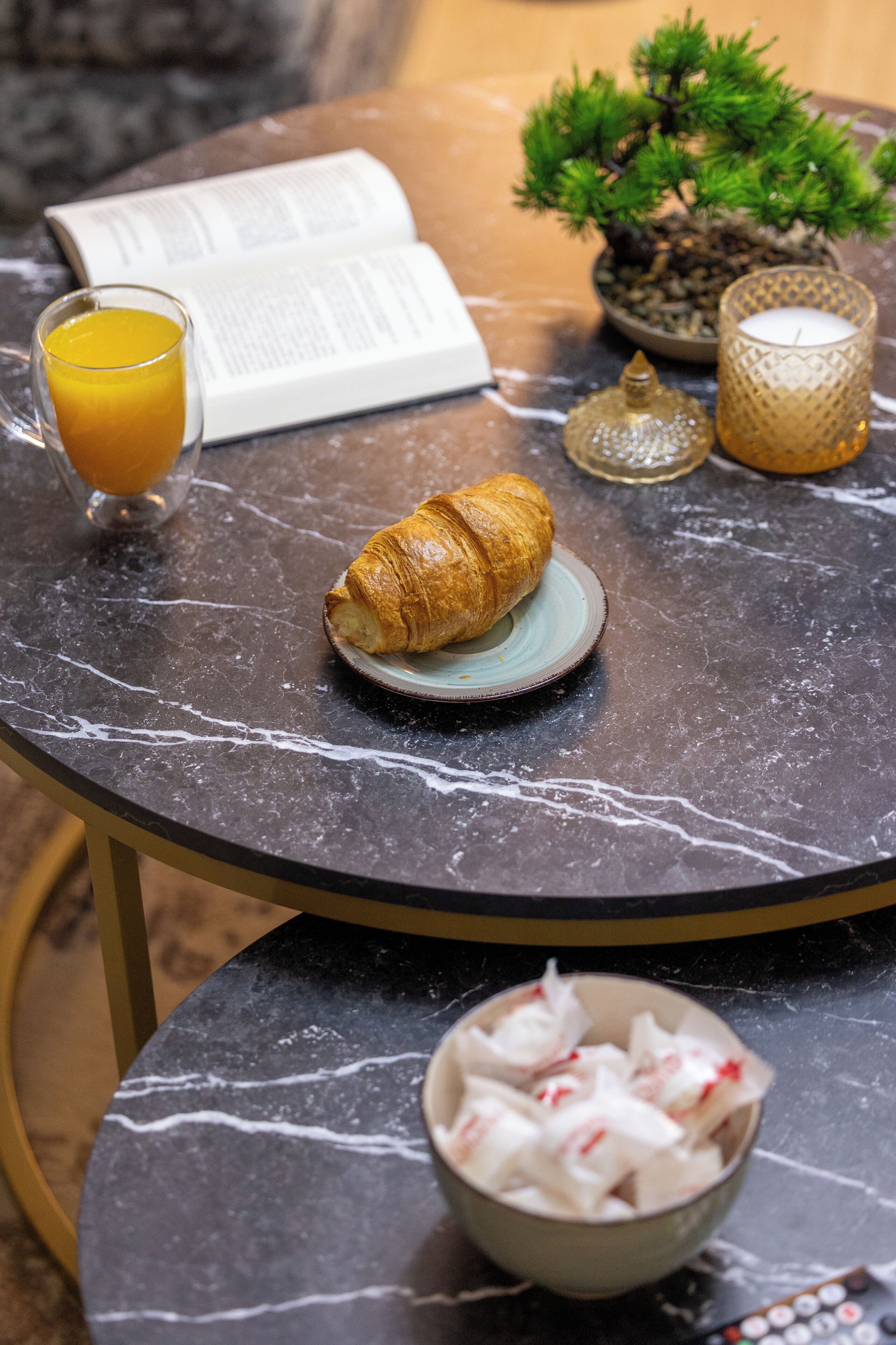 Une table ronde avec un livre, un verre de jus d’orange, un croissant, une bougie et un bonsaï.