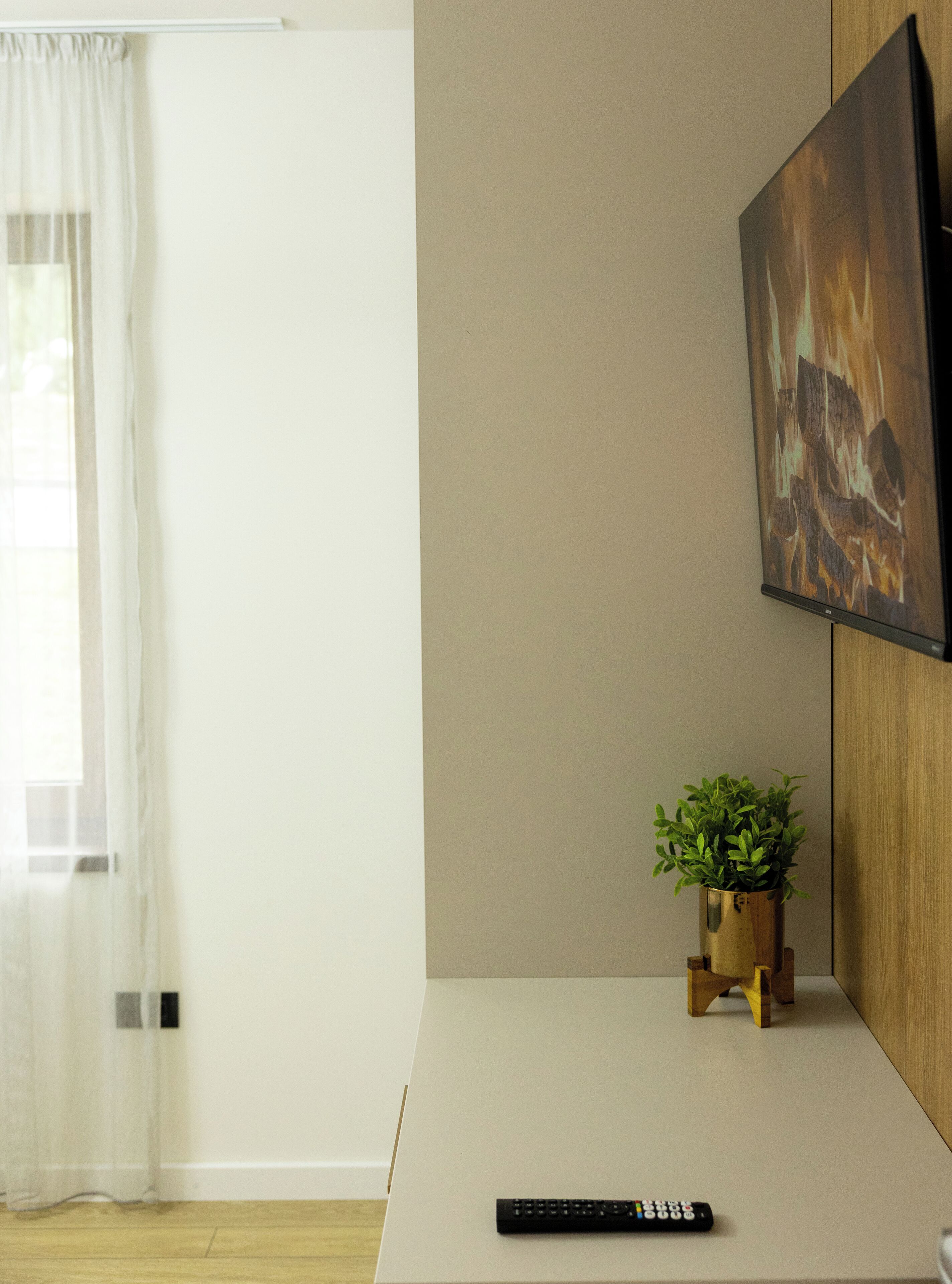 Un salon moderne avec un téléviseur à écran plat fixé au mur, une télécommande posée sur une surface blanche et une petite plante en pot sur un support en bois.