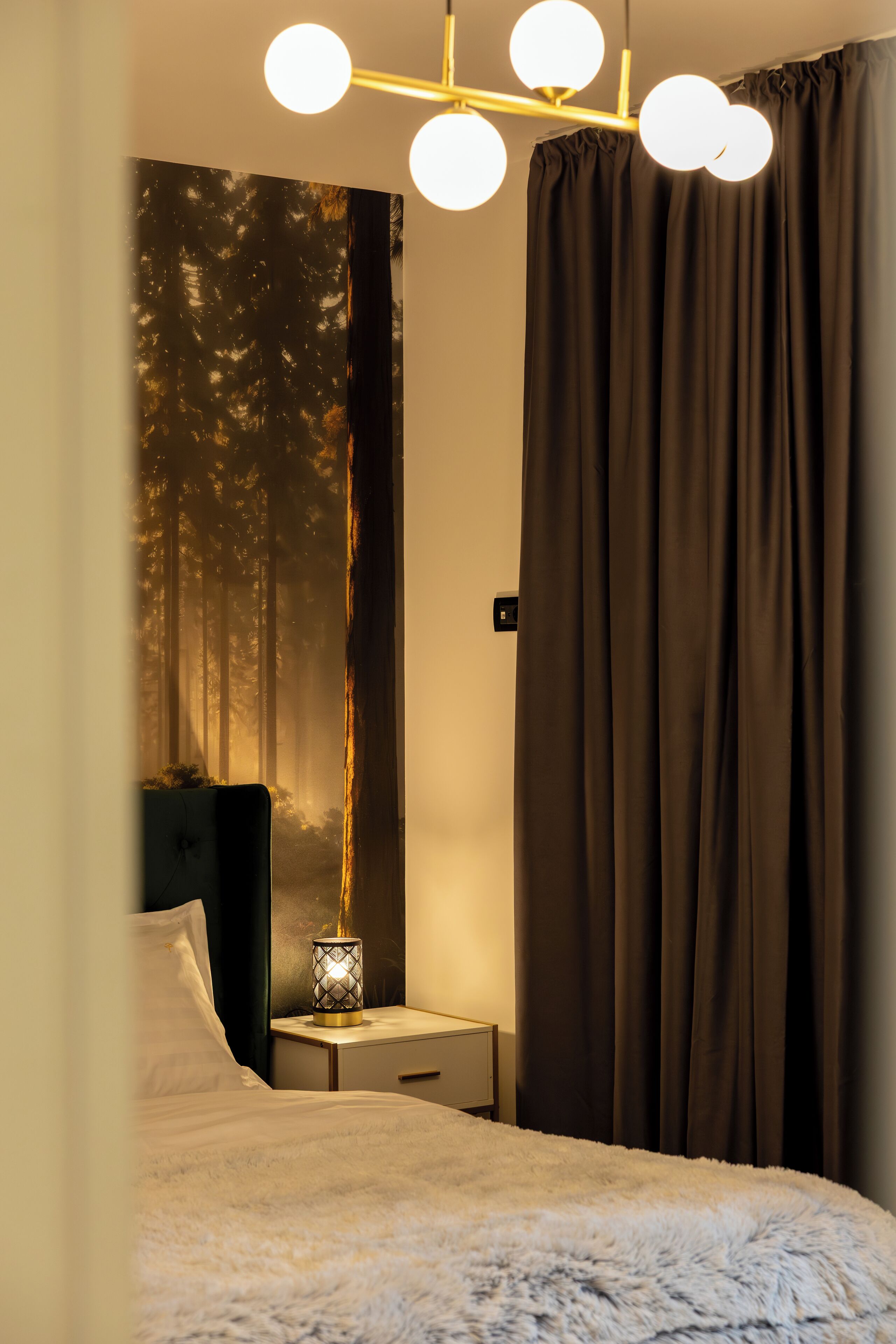 A bedroom with a bed, a nightstand, a wall-mounted lamp, and a large window with curtains.