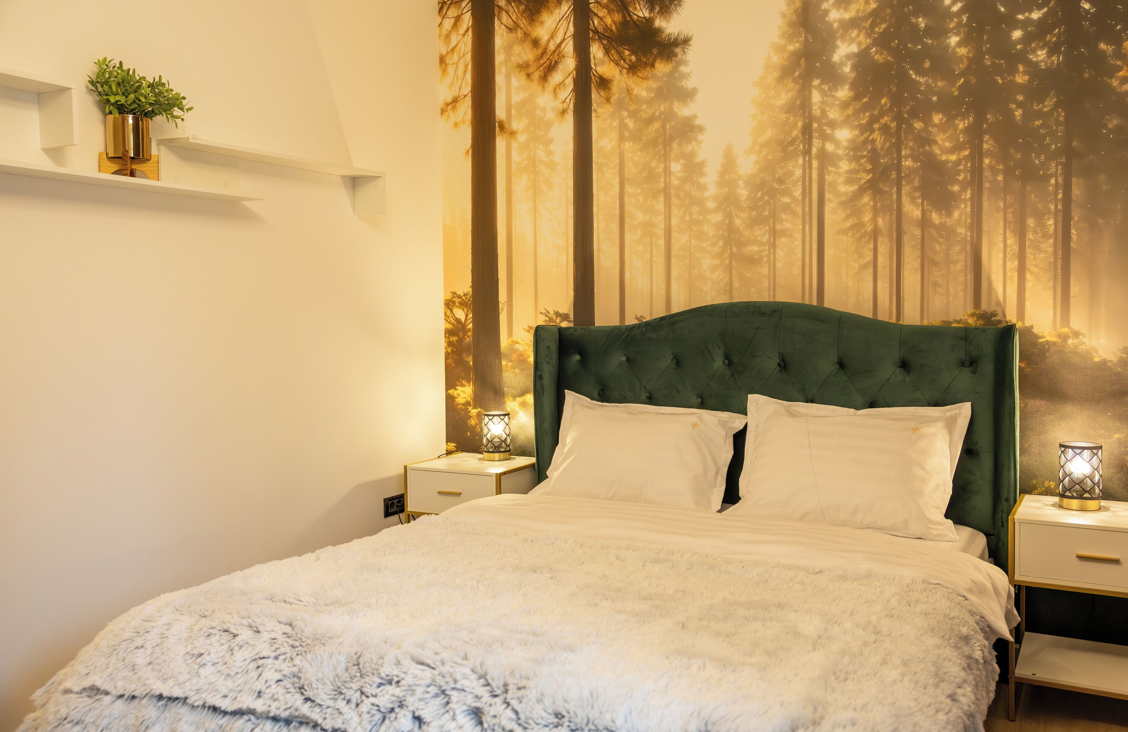 A bedroom with a large bed, white bedding, a green upholstered headboard, and a wall mural of a forest. There are bedside tables with lamps and a small plant on a shelf.