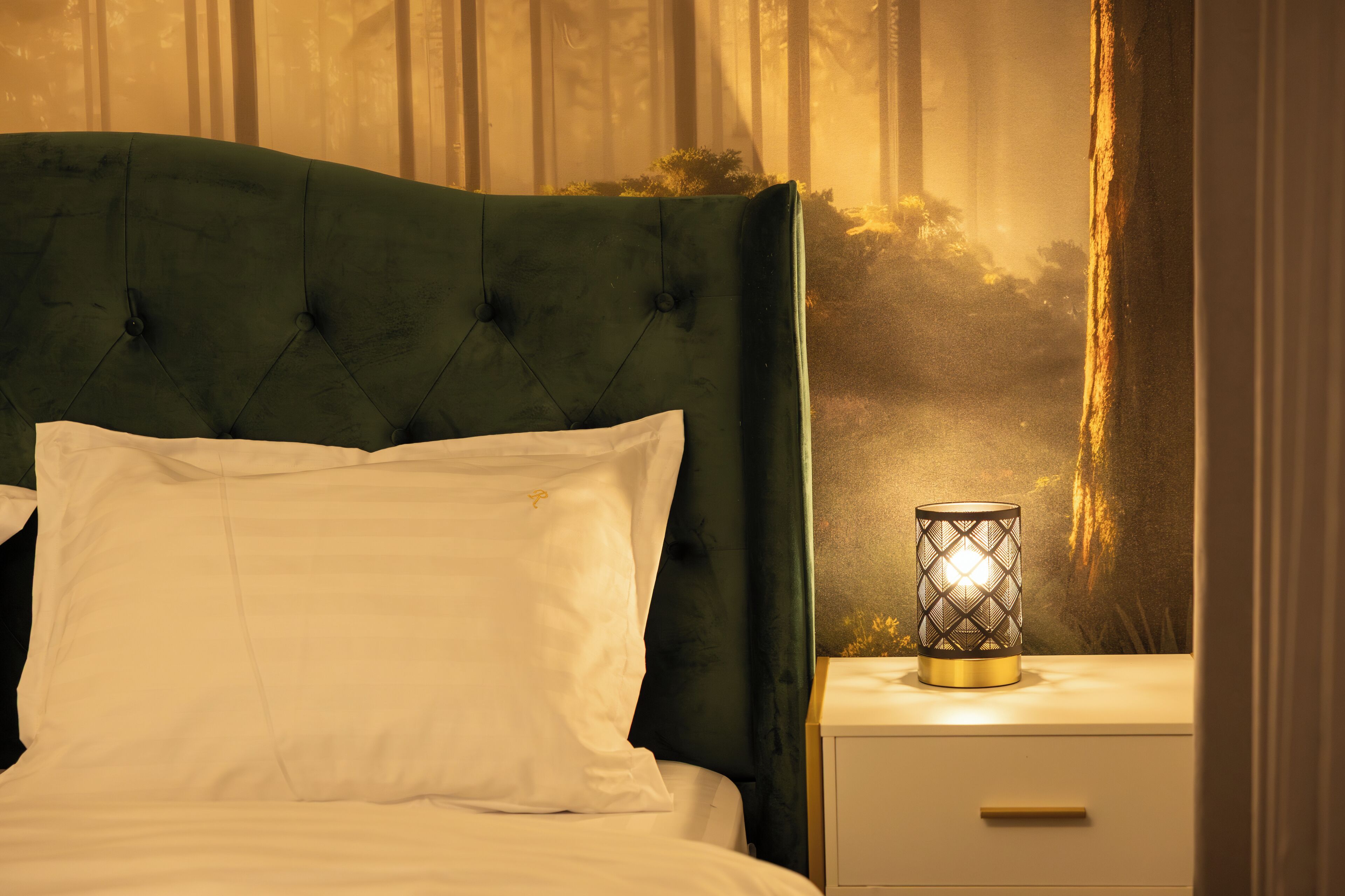 Una cama con cabecera tapizada de verde, almohadas blancas y una mesita de noche con una lámpara decorativa.
