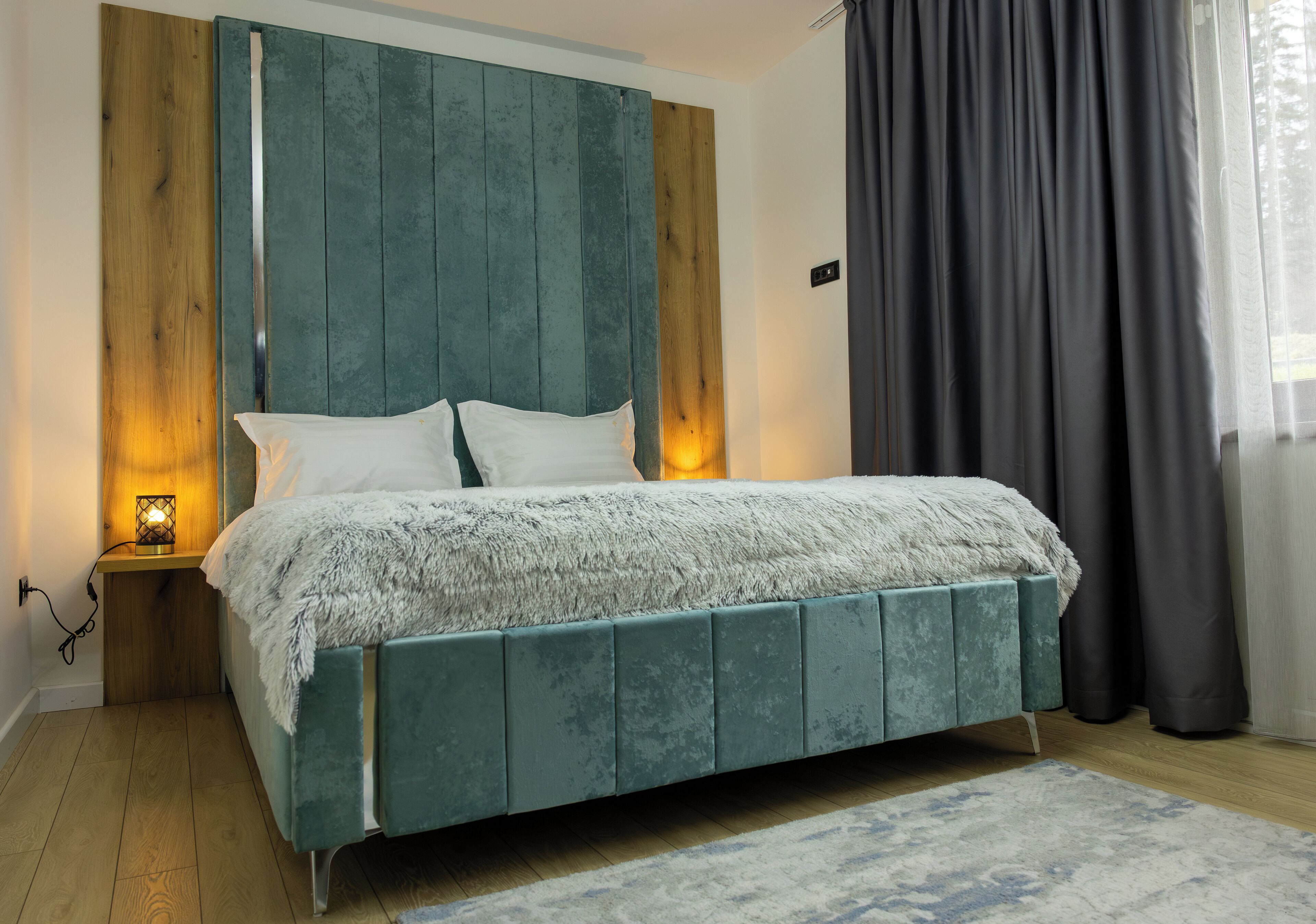 A bed with a teal headboard, white pillows, and a fluffy blanket. There is a small bedside table with a lamp.