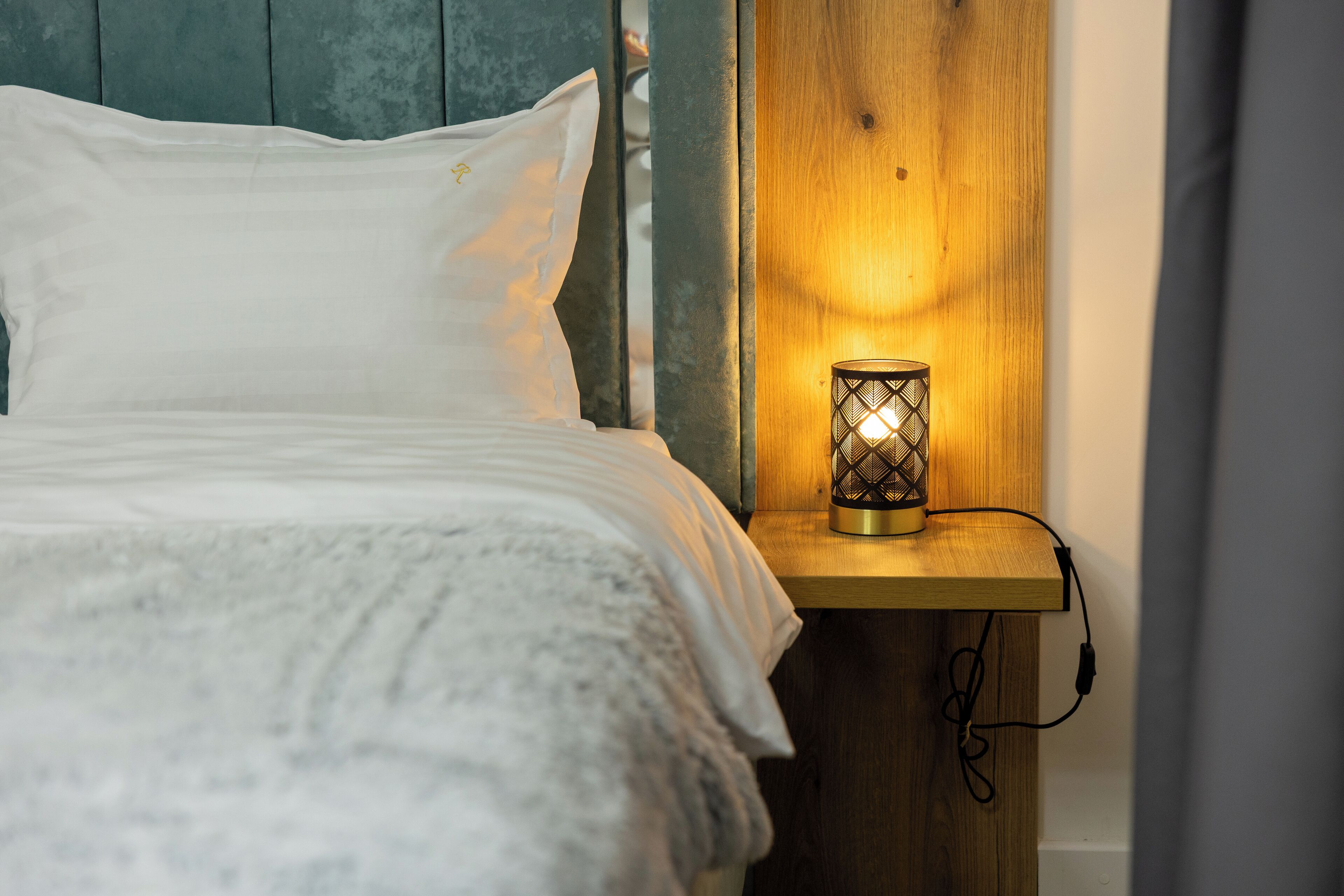 A bed with white linens, a wooden bedside table with a wall-mounted lamp, and a power cord.