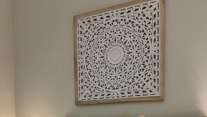 Een net opgemaakt bed met witte en mosterdkleurige kussens, een decoratieve wandhanger en opgerolde witte handdoeken op een beddengoed met dessin.