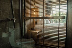 Une salle de bain moderne dotée d’une douche à parois de verre, de stores en bois et offrant une vue sur une chambre à coucher avec un lit et une table de chevet.