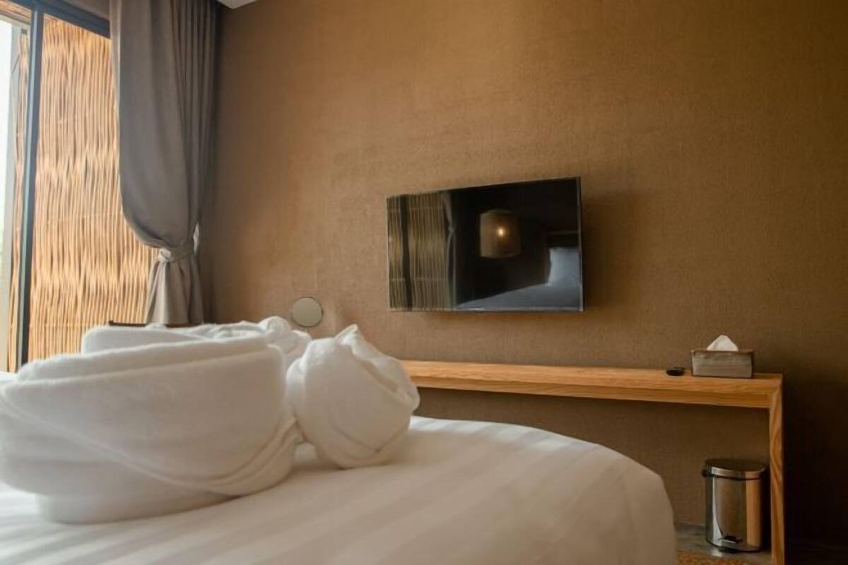 Una habitación de hotel con una cama, un televisor montado en la pared, una mesita de noche con caja de pañuelos y un cubo de basura.