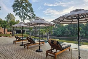 Une terrasse en bois, dotée de chaises longues et de parasols, qui offre une vue sur une étendue d’eau et sur la verdure.