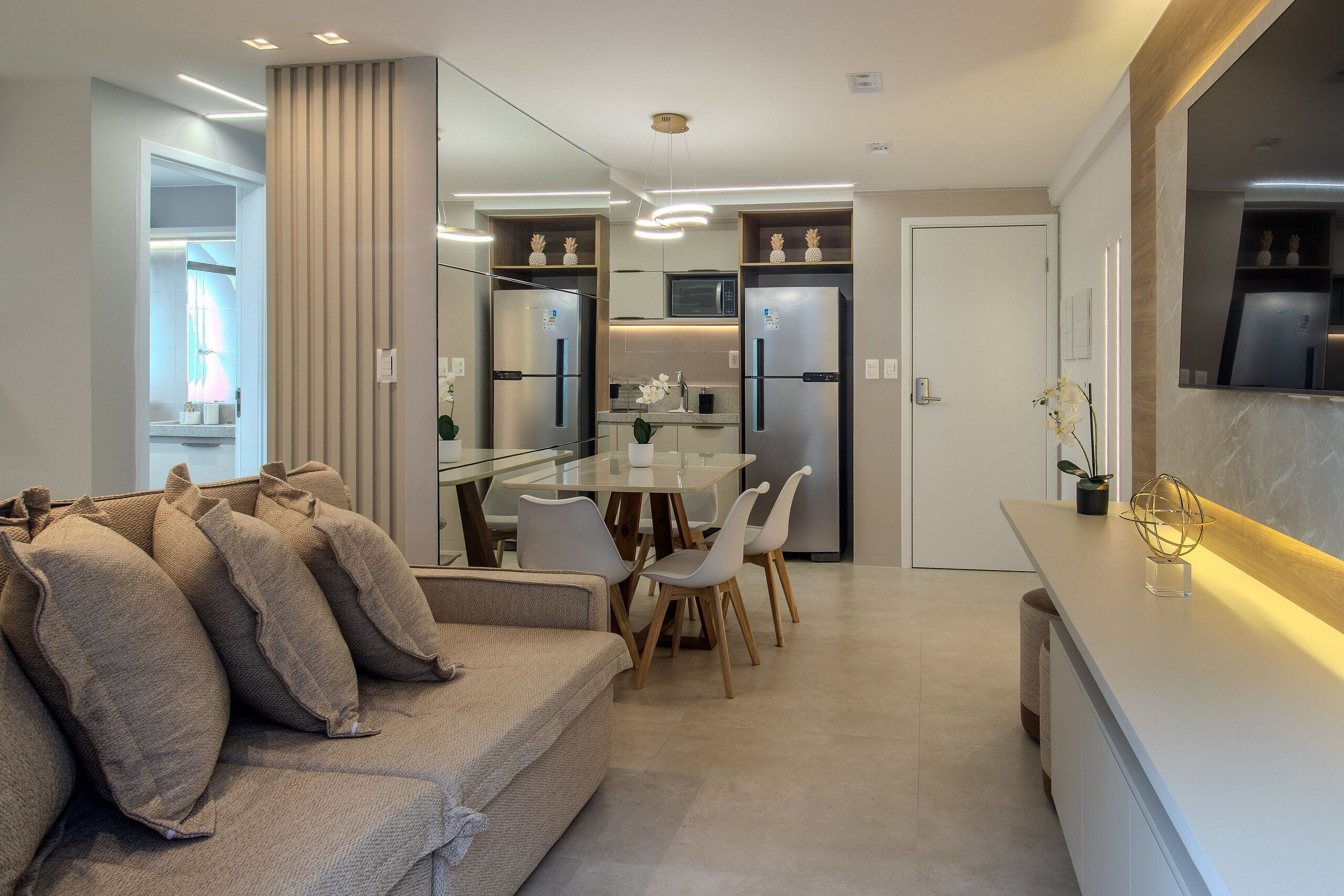 A modern kitchen with built-in appliances, a dining area with a glass table and chairs, and a living area with a sofa and cushions.