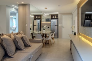 A modern kitchen with built-in appliances, a dining area with a glass table and chairs, and a living area with a sofa and cushions. - CSQ Jardins  (João Pessoa)