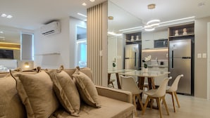 A modern living room with a sofa, dining table, chairs, and a kitchen area with appliances. - CSQ Jardins  (João Pessoa)