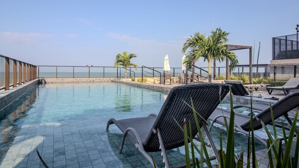 A rooftop pool with lounge chairs, palm trees, and a view of the sea. - CSQ Jardins  (João Pessoa)