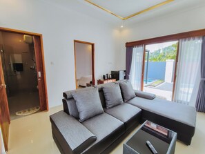 A modern living room with a grey sectional sofa, a coffee table, and a TV. There is a sliding glass door to a balcony with a pool view. - Bali Lovina Karuna Villa (kayu Putih)