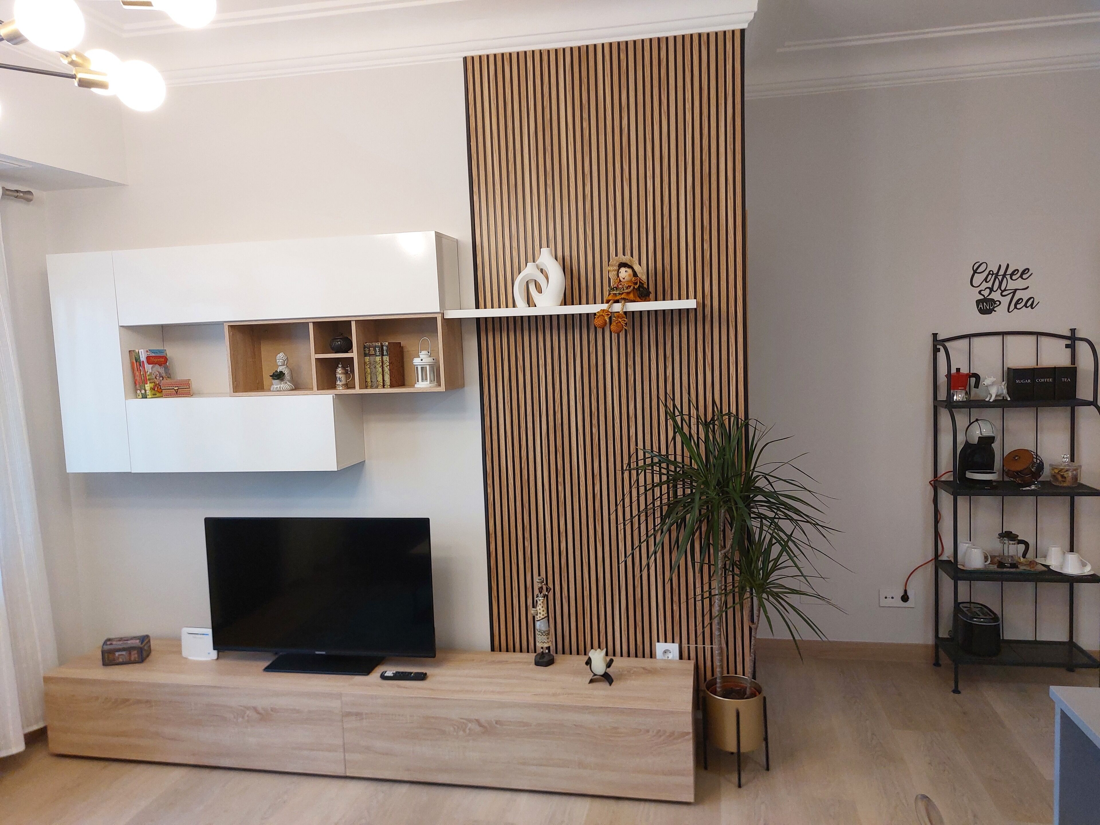 A modern living room with a flat-screen TV on a wooden console, a decorative shelf with a figurine, a vertical wooden panel, and a coffee and tea sign on the wall.