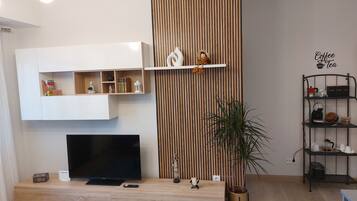 A modern living room with a flat-screen TV on a wooden console, a decorative shelf with a figurine, a vertical wooden panel, and a coffee and tea sign on the wall.