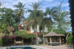 A pool area with a thatched gazebo, palm trees, and a house with a red roof. - Melati Resort & Hotel Kuta Lombok (Pujut)