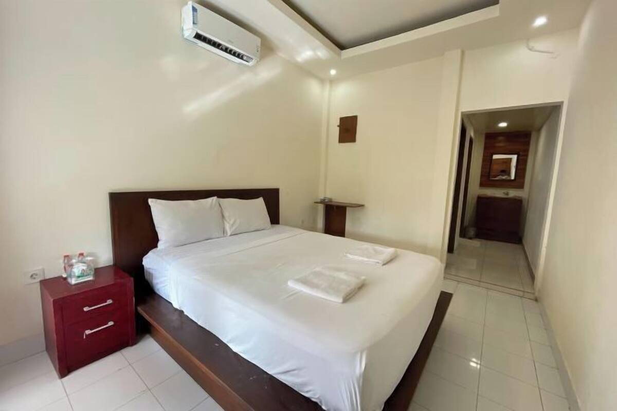 A hotel room with a bed, a nightstand, a red drawer, a white wall, and a ceiling-mounted air conditioner.
