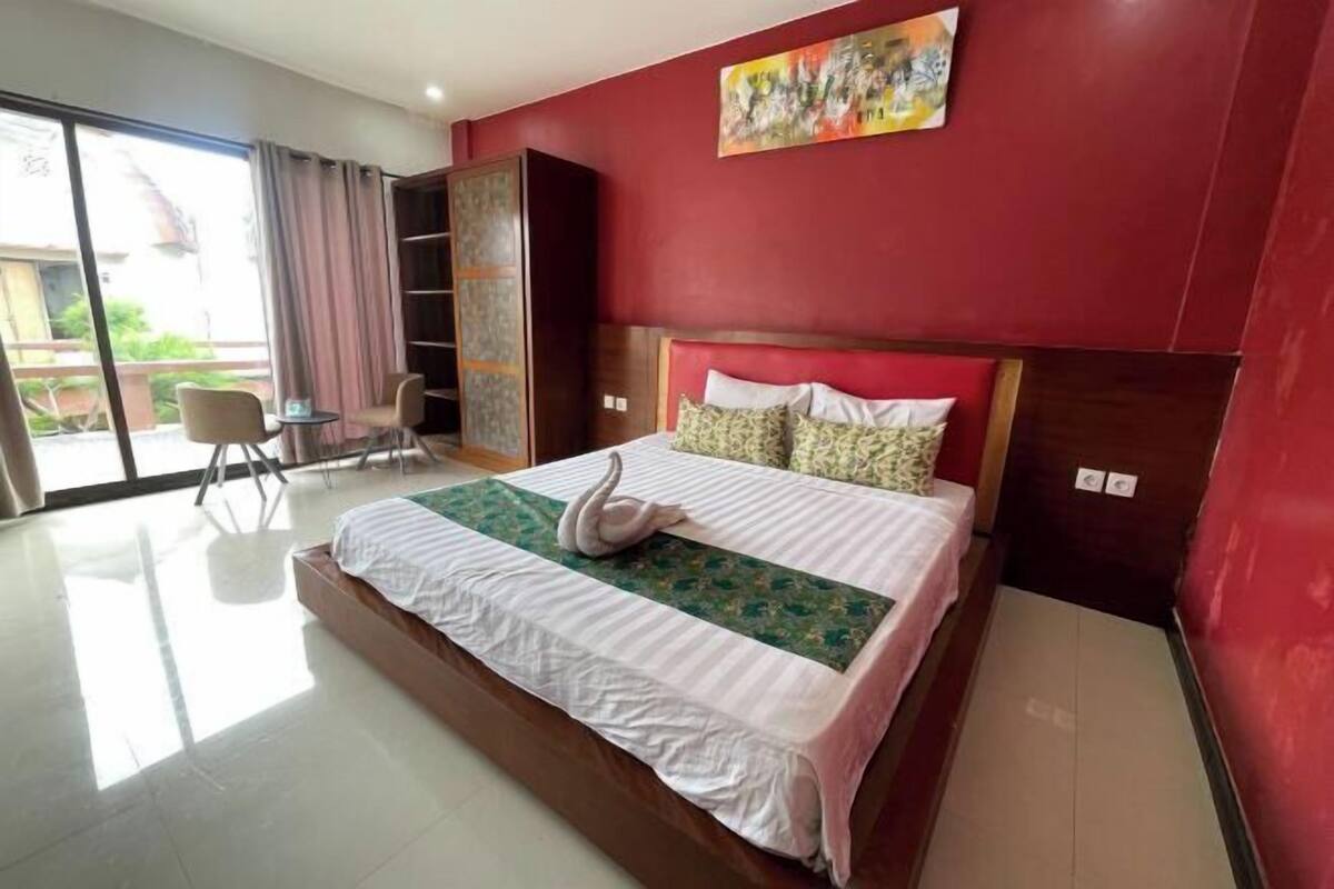 A hotel room with a large bed, a wooden headboard, a green and white patterned bedspread, a swan decoration, a painting on the wall, a sliding glass door, and a small table with chairs.