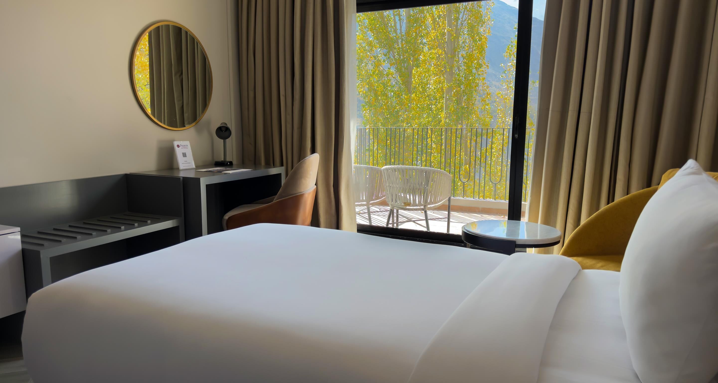Camera d'albergo con un letto, una scrivania, una sedia, un tavolino, uno specchio e vista su alberi e un balcone.