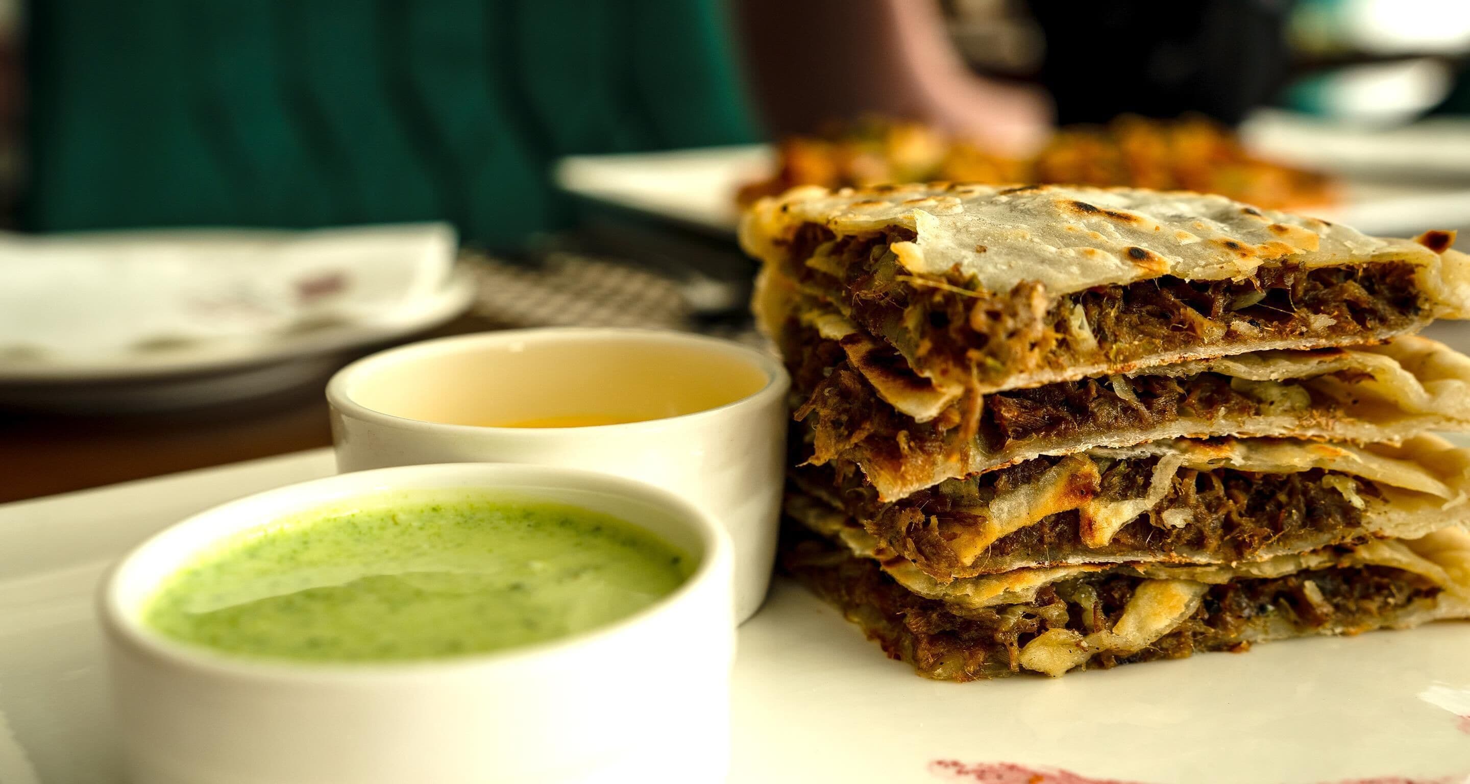 A stack of layered food items with two small bowls of green and yellow sauces.
