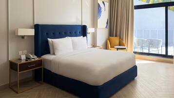 Une chambre d’hôtel avec un grand lit, une table de chevet avec un téléphone et une lampe, une chaise, et un balcon avec des meubles d’extérieur.