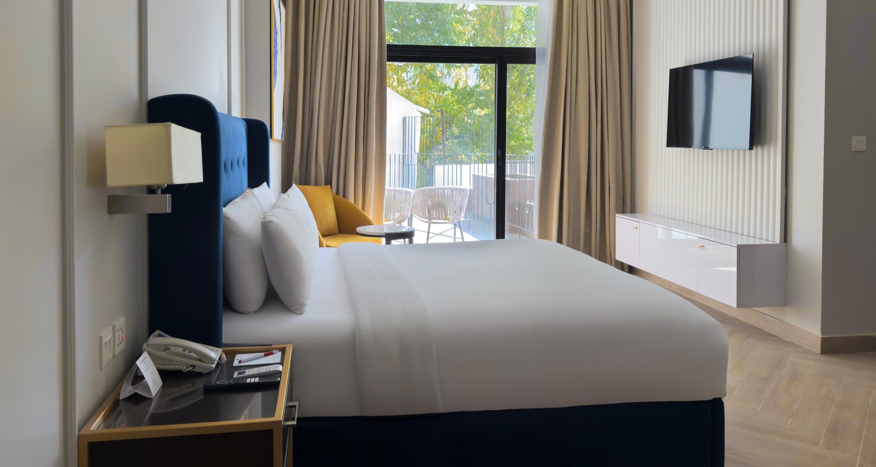 Quarto de hotel com cama grande, mesa de cabeceira com luminária, TV fixada na parede e vista para o exterior através de uma porta deslizante.