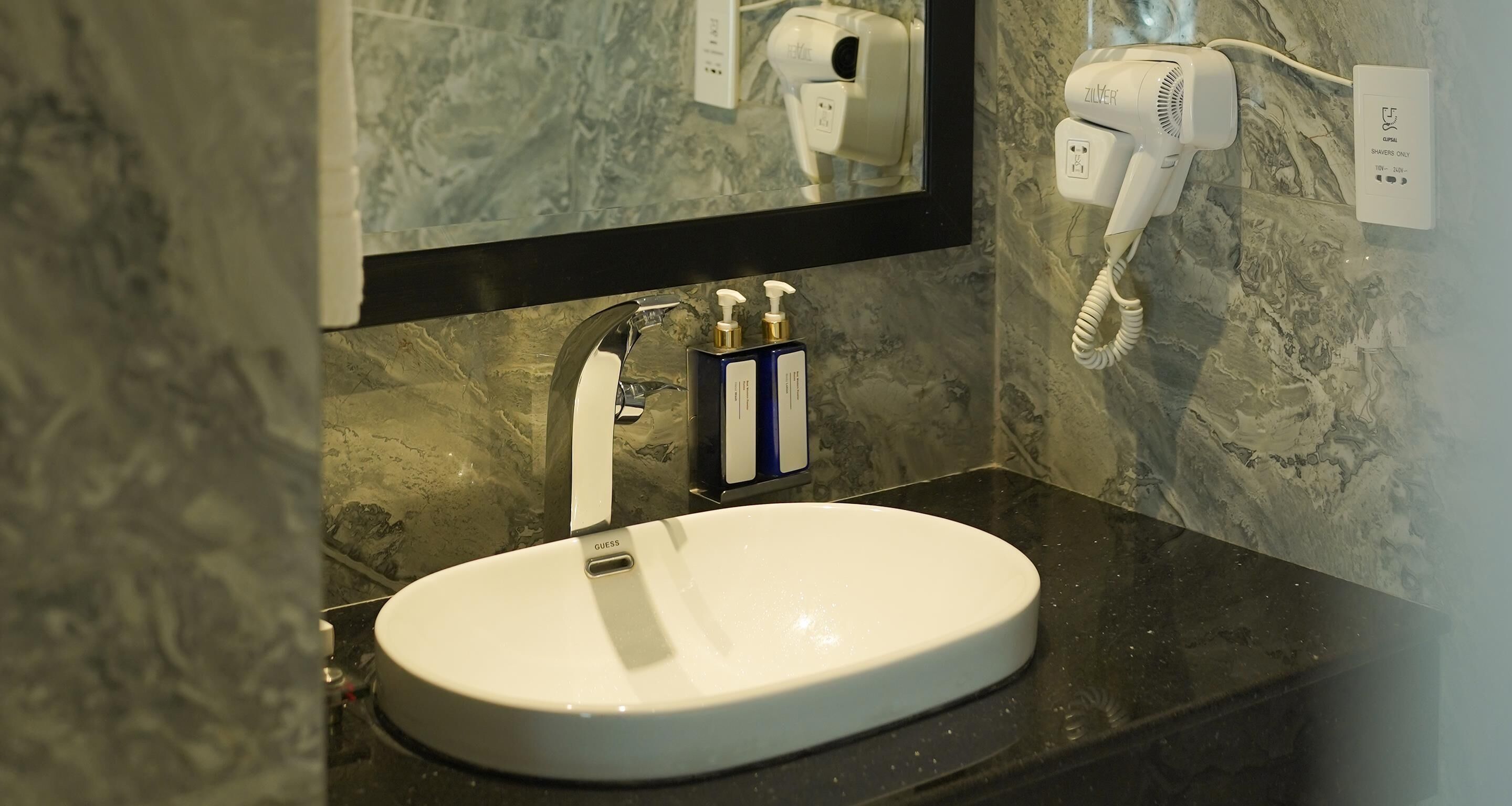 Un baño con lavamanos blanco, un espejo, un secador de pelo y un enchufe en la pared.