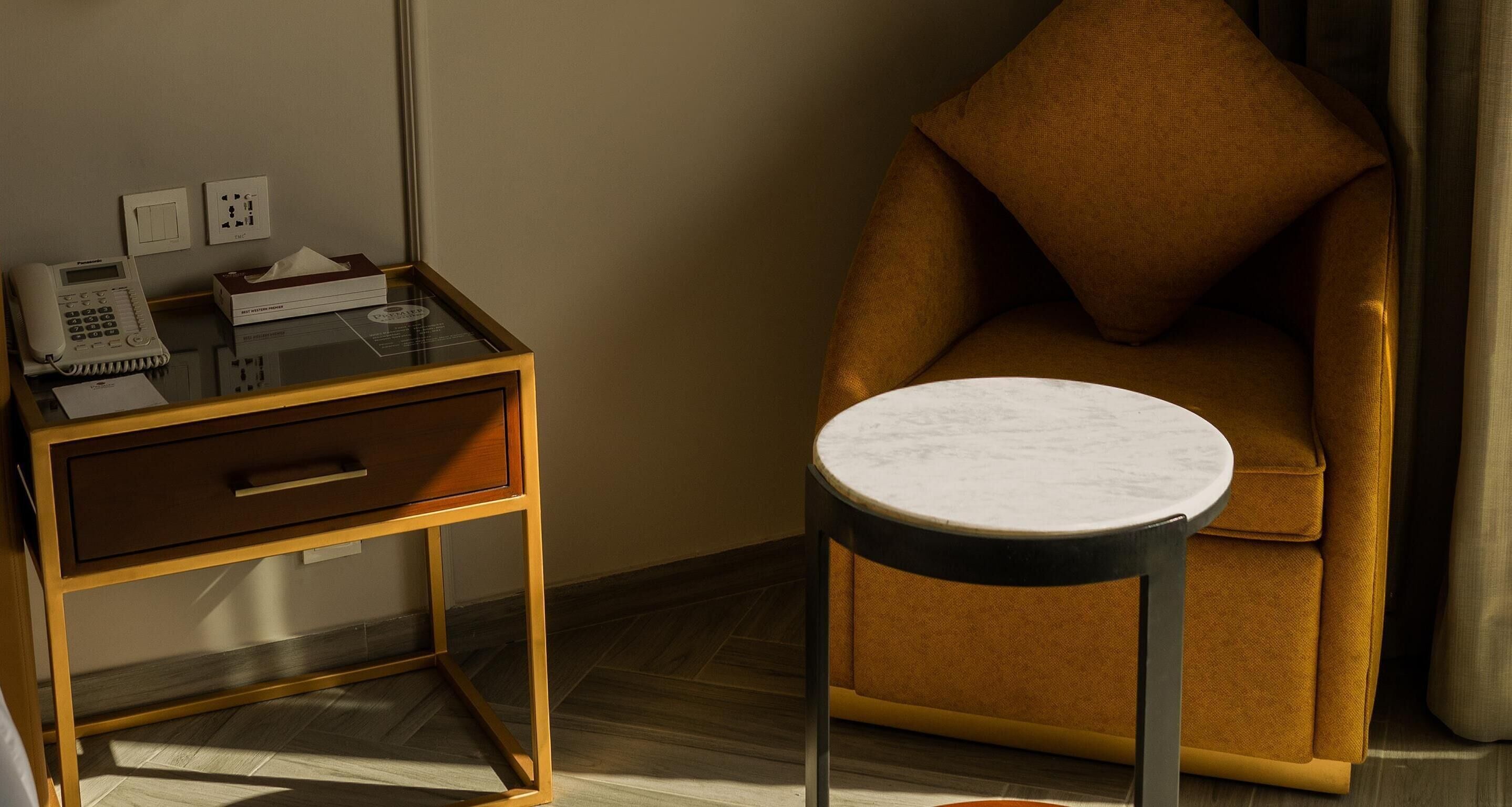 Una habitación de hotel con una mesita de noche de madera, un teléfono, una caja de pañuelos y una pequeña mesa redonda con tapa de mármol.