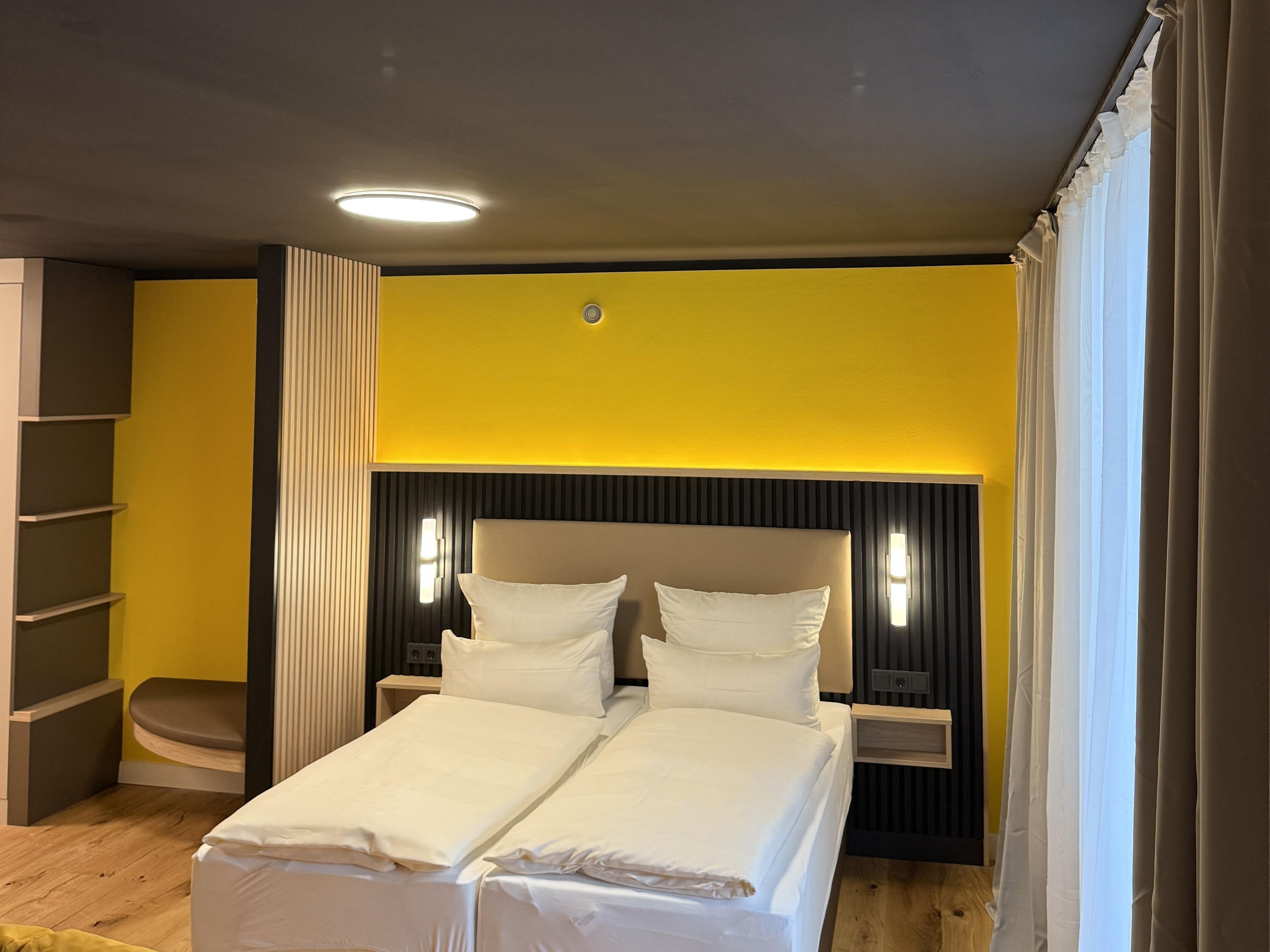 Una habitación de hotel con cama, mesitas de noche, estantería y ventana con cortinas.