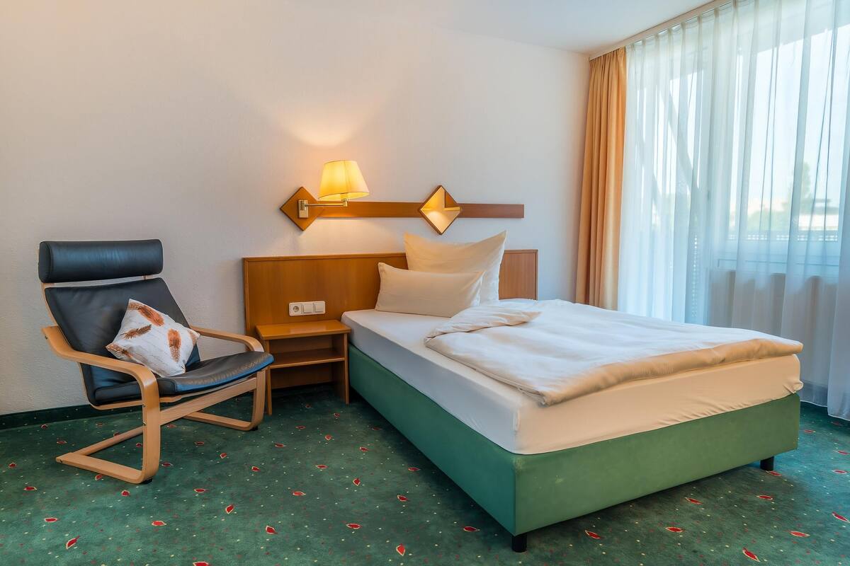 Une chambre d’hôtel avec un lit, une chaise, une table de chevet et une lampe fixée au mur.