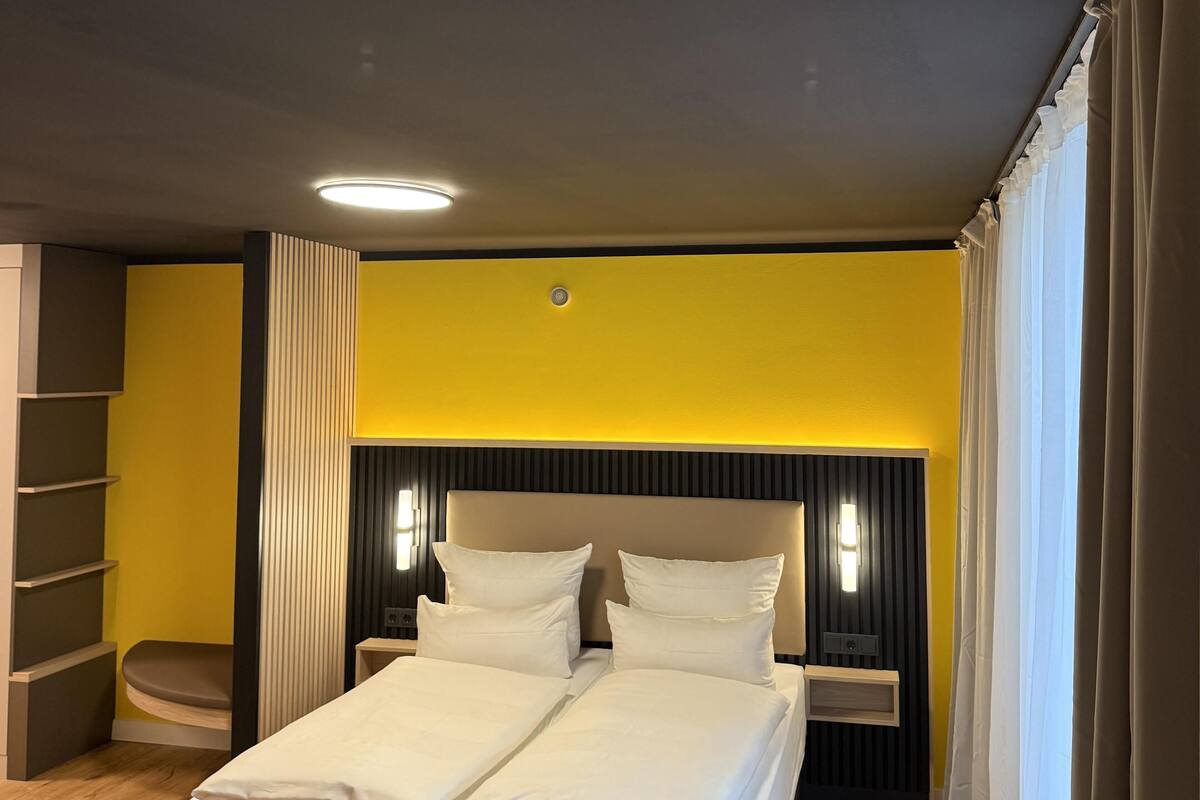 Une chambre d’hôtel comprenant un lit, des tables de chevet, une petite table, une lampe fixée au mur et une fenêtre avec des rideaux.