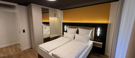 Una cama doble con ropa de cama blanca, suelo de madera, una lámpara de techo y una mesita de noche fijada a la pared.