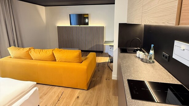 A modern hotel room with a yellow sofa, a wooden floor, a built-in kitchen with a stove, and a large window. - Residenzhotel Stuttgart Airport, Sure Hotel Collection by BW (Steinenbronn)