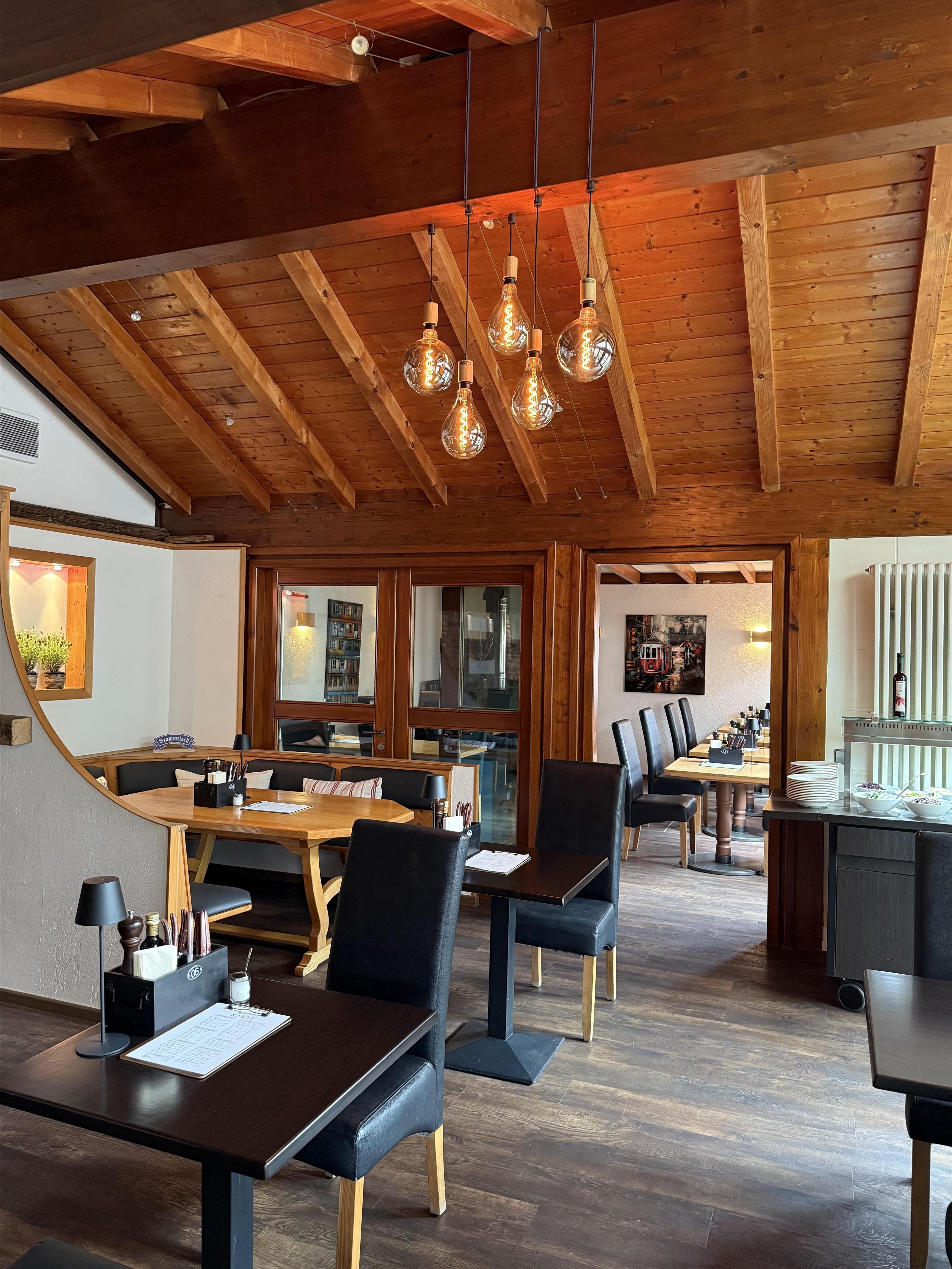 A restaurant with wooden beams, hanging light fixtures, and a mix of wooden and upholstered tables and chairs.