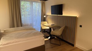 Quarto de hotel moderno com cama, escrivaninha, poltrona, televisão e janela com cortinas.