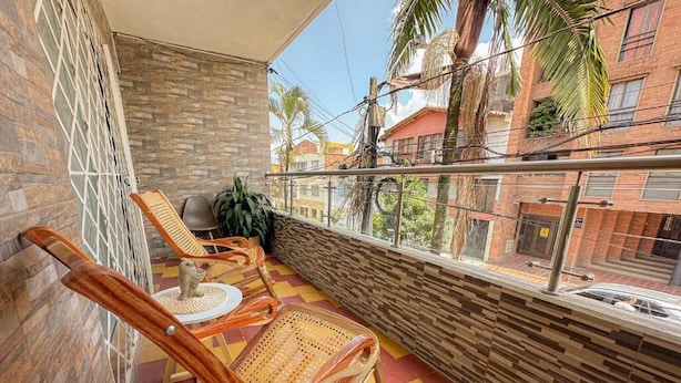 Un balcon doté d’un sol à carreaux, de deux fauteuils en osier et offrant une vue sur des immeubles et des palmiers.