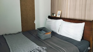 A neatly made bed with a grey blanket, a grey storage bag, and a wooden headboard with a small shelf holding a plant and a picture frame. - Mall of Asia - Cheap Accommodations (Pasay)