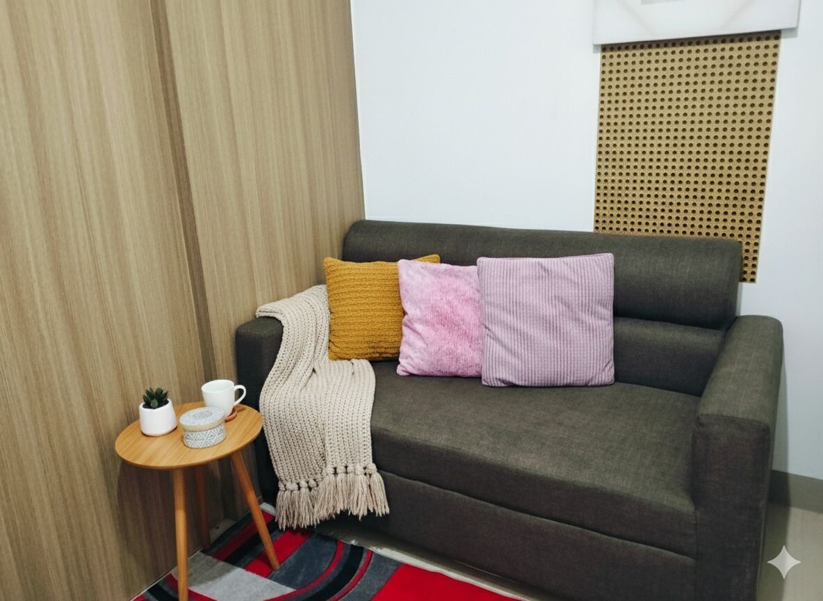 Un sofá gris con una manta beige y dos almohadas, una mesita de madera con una taza y una planta, y una alfombra a rayas rojas y negras.